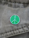 A small circular enamel pin featuring a green peace sign with a silver border, shown pinned to a gray woven fabric surface.