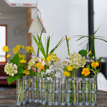 A decorative arrangement of mixed wildflowers in clear glass test tubes on a rustic wooden table, with a softly blurred indoor background and a hanging pendant light.