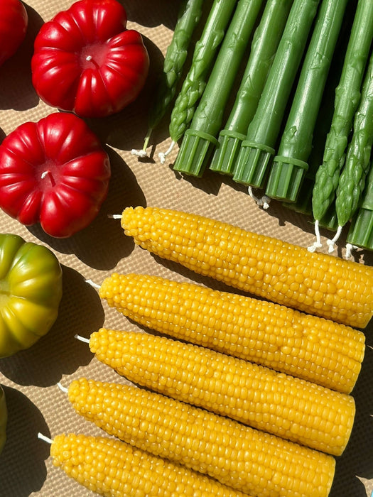 A styled flat-lay of novelty candles shaped like whole corn cobs, green asparagus spears, red and green tomatoes, and a brown woven mat underneath.