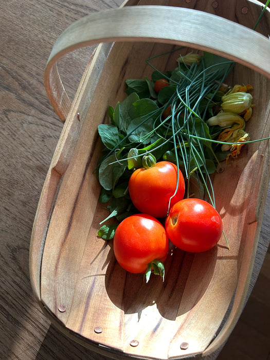 A wooden basket with a curved handle holds fresh produce, including three red tomatoes, green herbs, and yellow flowers, photographed on a wooden tabletop.