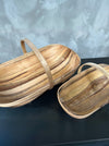 Two handcrafted woven wooden baskets with curved handles, displayed on a dark surface against a textured gray background.