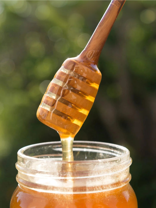 A wooden honey dipper dripping golden honey into a clear glass jar, with a soft blurred green outdoor background.