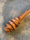 A close-up of a carved wooden spoon or honey dipper with a spiral ridged head, made from olive wood with a warm brown and tan grain, resting on a textured gray concrete surface.