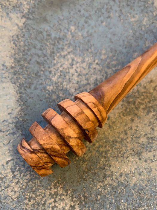 A close-up of a carved wooden spoon or honey dipper with a spiral ridged head, made from olive wood with a warm brown and tan grain, resting on a textured gray concrete surface.