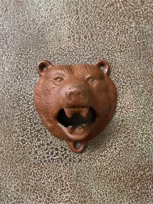 A rustic cast iron bear head bottle opener with a textured brown finish, shown against a matching textured metallic background.