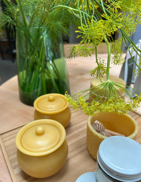 Two round yellow beeswax honey pots with lids sit on a wooden board next to a glass jar of honey and a wooden honey dipper, with a green glass vase of dill in the background.