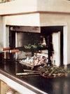 An outdoor wood-burning pizza oven with a dark countertop in the foreground holding tongs, herbs, and a tray of toppings, set against a light-colored stone wall.