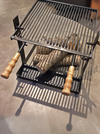 A black metal fire pit grill with a grate top, lower tray, and two turned wooden handles, shown with stacked firewood logs inside on a concrete floor.