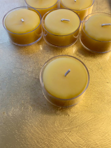 Five small beeswax tea lights in clear glass holders arranged on a textured gold surface.
