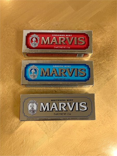 Three Marvis toothpaste tubes stacked vertically on a gold metallic surface. The top tube is Cinnamon Mint with a red label, the middle is Aquatic Mint with a blue label, and the bottom is Whitening Mint with a silver label.