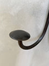 A close-up of a dark metal wall hook with a rounded, mushroom-like top and a curved stem, mounted against a textured off-white wall.