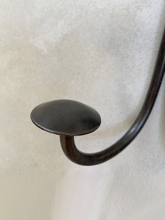 A close-up of a dark metal wall hook with a rounded, mushroom-like top and a curved stem, mounted against a textured off-white wall.