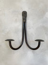 A rustic black metal double hook wall hanging with a curved U-shape design and flat circular end caps, mounted on a textured light gray plaster wall.