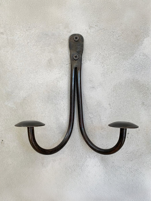 A rustic black metal double hook wall hanging with a curved U-shape design and flat circular end caps, mounted on a textured light gray plaster wall.
