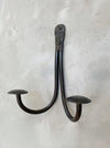 A rustic black metal double hook wall hanger with mushroom-shaped ends, mounted on a textured white plaster wall.