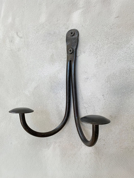 A rustic black metal double hook wall hanger with mushroom-shaped ends, mounted on a textured white plaster wall.