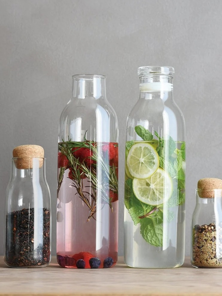 Three clear glass bottles with cork stoppers sit on a light wood surface against a gray wall. The center-left bottle contains pink liquid with rosemary and berries, the center-right bottle contains clear liquid with lime slices and mint, and the side bottles contain dark and light seeds or spices.
