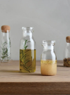 Two clear glass bottles with cork stoppers sit on a wooden surface against a soft gray background. The taller bottle contains golden oil with a green herb sprig inside, and the shorter bottle holds a pale yellow liquid with small specks.