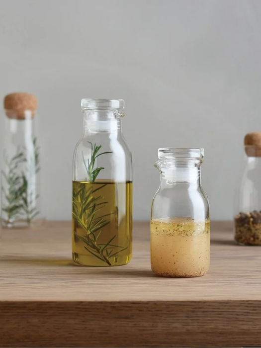 Two clear glass bottles with cork stoppers sit on a wooden surface against a soft gray background. The taller bottle contains golden oil with a green herb sprig inside, and the shorter bottle holds a pale yellow liquid with small specks.