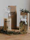 Two clear glass bottles with white stoppers and kraft paper labels reading BOTTLIT, arranged on a wooden surface with rosemary sprigs and a brown box in the background.
