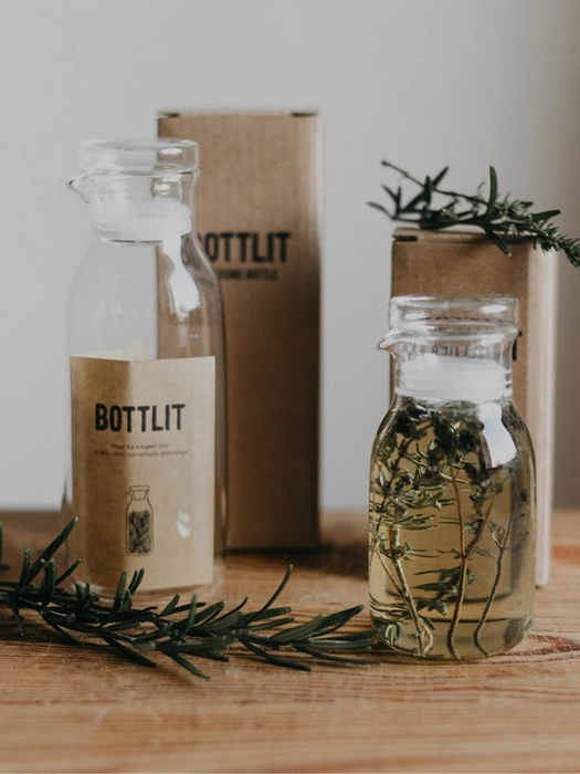 Two clear glass bottles with white stoppers and kraft paper labels reading BOTTLIT, arranged on a wooden surface with rosemary sprigs and a brown box in the background.