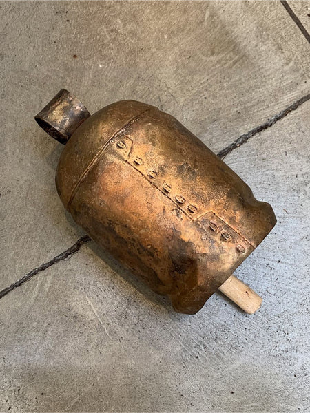 A rustic antique-style bronze bell with a wooden mallet handle, resting on a gray concrete floor.