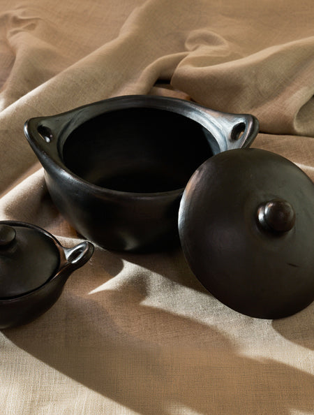 A dark matte ceramic pot with its matching lid resting beside it on draped beige fabric. A smaller matching vessel is partially visible in the lower left corner.