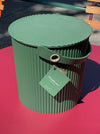 A green ribbed metal storage bin with a matching lid and handle, featuring a hanging tag, photographed outdoors on a bright pink surface with blurred park furniture in the background.
