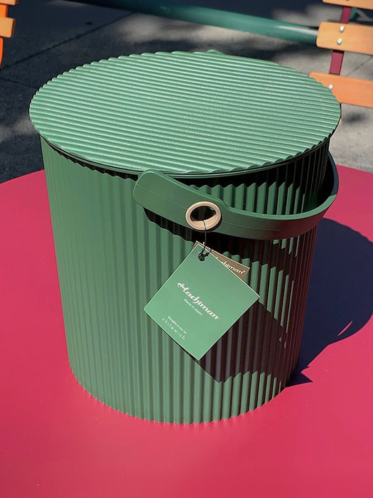 A green ribbed metal storage bin with a matching lid and handle, featuring a hanging tag, photographed outdoors on a bright pink surface with blurred park furniture in the background.