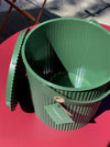 A green ribbed plastic bucket with a matching lid, resting on a bright pink-red table outdoors under direct sunlight. A hangtag is attached to the rim.