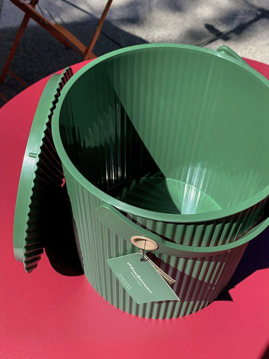 A green ribbed plastic bucket with a matching lid, resting on a bright pink-red table outdoors under direct sunlight. A hangtag is attached to the rim.