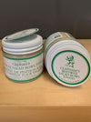 Two jars of Clapham's Beewax Salad Bowl Finish shown on a light wood surface against a dark gray background. One jar is upright and the other is lying on its side, both with white lids and labels featuring green borders and text.