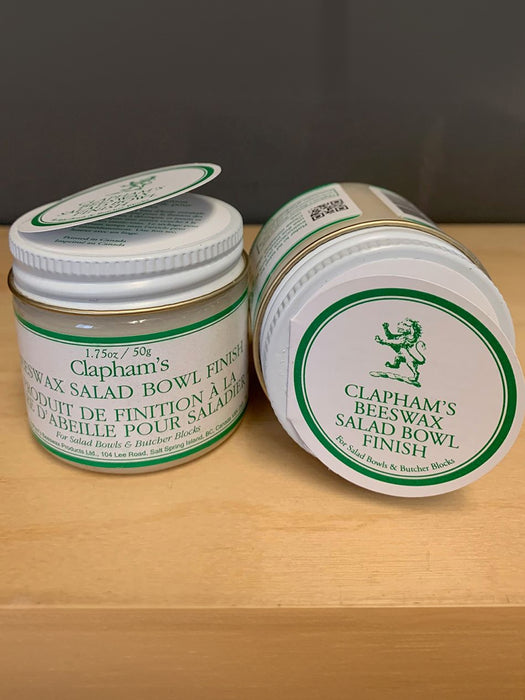 Two jars of Clapham's Beewax Salad Bowl Finish shown on a light wood surface against a dark gray background. One jar is upright and the other is lying on its side, both with white lids and labels featuring green borders and text.