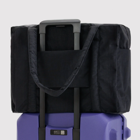 A black soft-sided duffel bag with a telescoping handle, shown resting on top of a purple rolling suitcase against a light gray studio background.