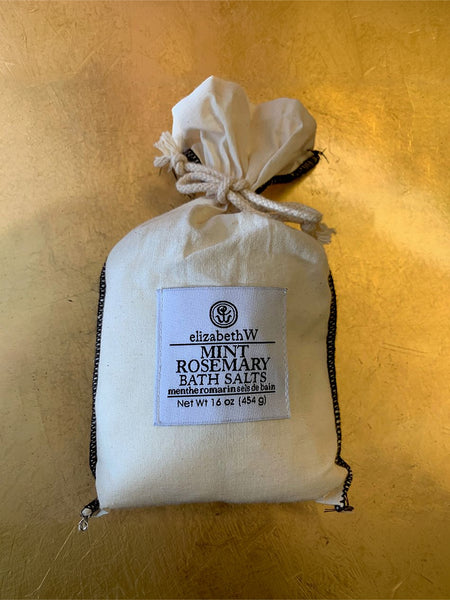 A drawstring pouch of ElizabethW Mint Rosemary Bath Salts, made of off-white canvas with black trim and a white label, set against a textured gold background.