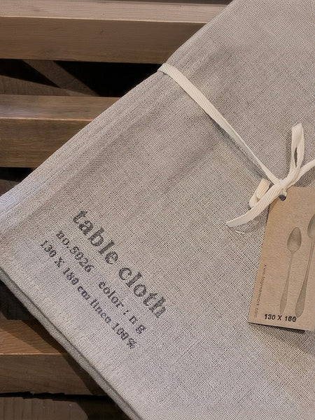 A folded light beige linen tablecloth tied with a cream ribbon, shown with a paper hangtag on a wooden surface. The cloth is printed with 'table cloth' and product details.