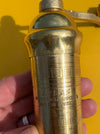 A close-up of a hand holding a brass Atlas pepper mill against a bright yellow background. The cylindrical mill has engraved Greek characters and the text 'ATLAS PEPPER MILL IMPORTS MADE IN GREECE'.
