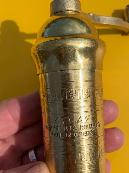 A close-up of a hand holding a brass Atlas pepper mill against a bright yellow background. The cylindrical mill has engraved Greek characters and the text 'ATLAS PEPPER MILL IMPORTS MADE IN GREECE'.
