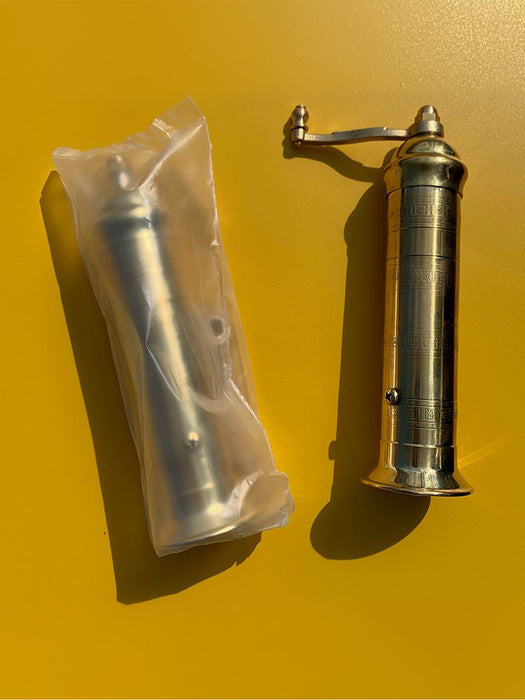 A brass hand-crank grinder shown next to its clear plastic packaging on a solid yellow background.