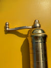 A close-up of a polished silver metal pepper mill grinder with a hand-crank handle, set against a vibrant yellow background.