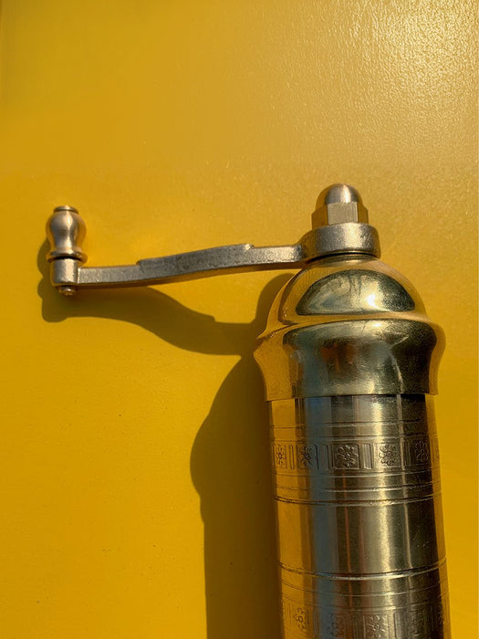 A close-up of a polished silver metal pepper mill grinder with a hand-crank handle, set against a vibrant yellow background.