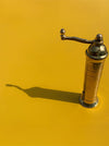 A vintage brass coffee grinder with a curved handle, standing on a vibrant yellow surface that casts a long shadow to the left.