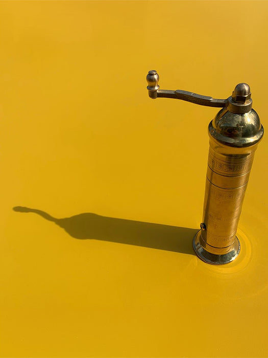A vintage brass coffee grinder with a curved handle, standing on a vibrant yellow surface that casts a long shadow to the left.