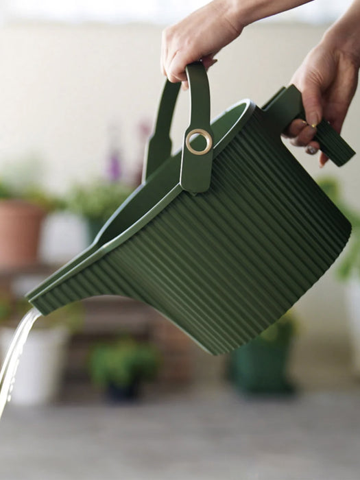 A green ribbed plastic watering bucket with a handle, being held by hands as water drains from the spout in a softly blurred indoor setting with potted plants.