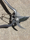 A close-up of pruning shears with dark gray metal handles and blades, resting on a textured concrete surface with a stone under the pivot point.