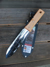 A large fixed-blade knife with a silver blade featuring depth markings and a light wood handle, resting on its black leather sheath over a dark wooden plank surface.