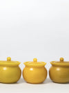 Three small round lidded jars in a matte mustard yellow color, arranged in a row against a plain white background.
