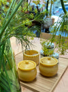 Three small yellow beeswax jars with lids, including one textured honey pot and two solid pots, arranged on a wooden tray next to a glass vase of dill in a cafe setting.