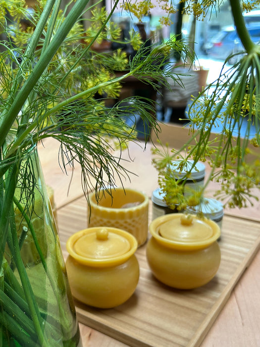 Three small yellow beeswax jars with lids, including one textured honey pot and two solid pots, arranged on a wooden tray next to a glass vase of dill in a cafe setting.