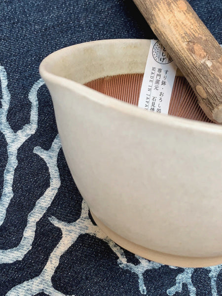 A close-up of a handmade ceramic bowl in a warm off-white glaze, resting on a navy blue patterned fabric. A wooden pestle and a paper tag with Japanese text are visible inside the bowl.